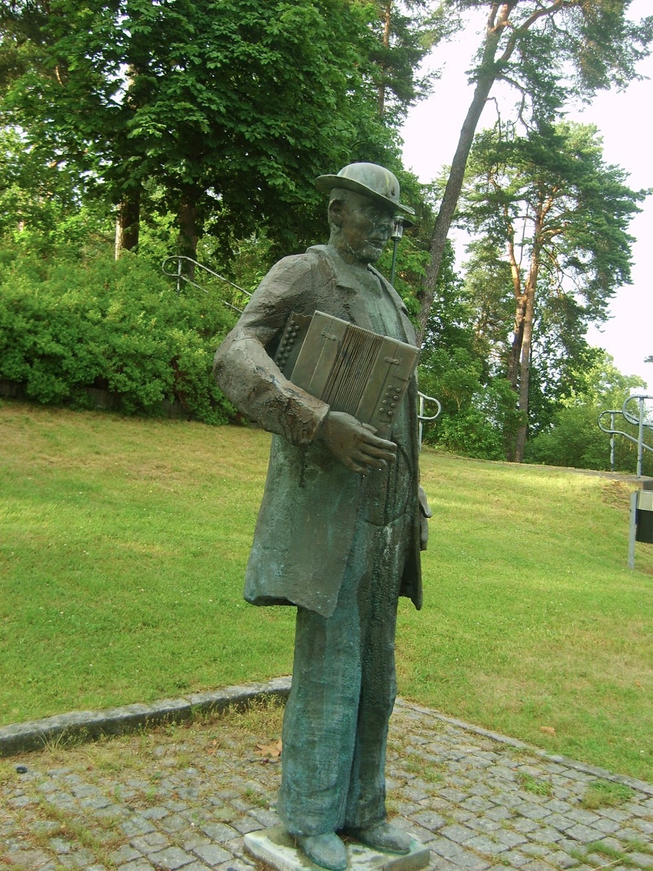Bronsstaty av en svensk spelman med dragspel i en park.